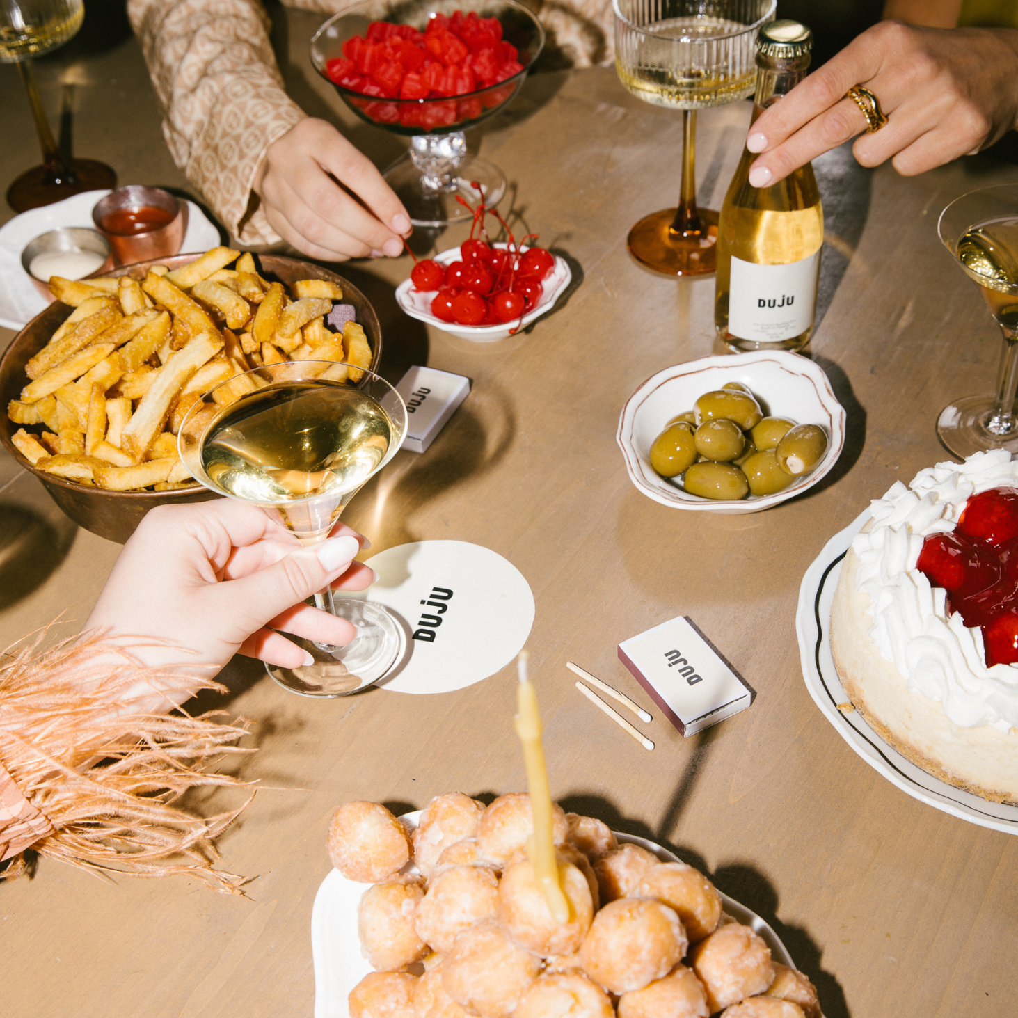 Table with food and drinks, including fries, olives, and a cake, with people around.