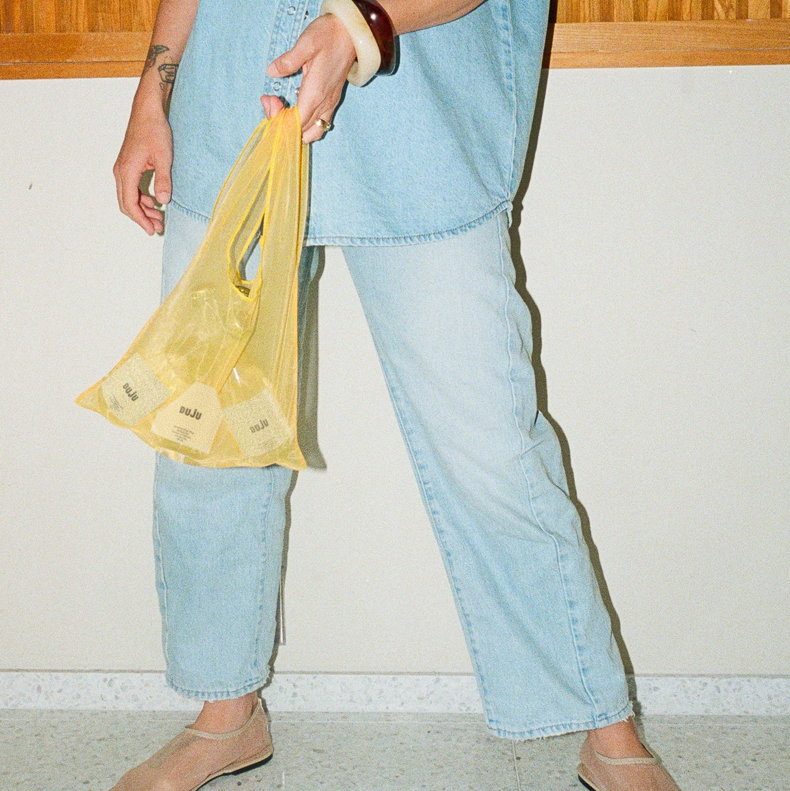 Person wearing a light blue denim outfit holding a yellow plastic bag indoors.