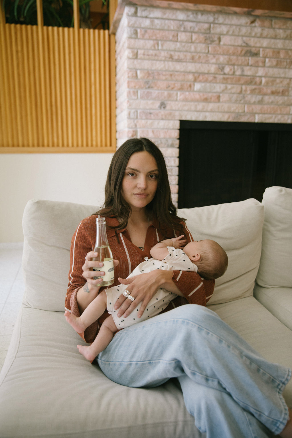 Woman holding a baby and a bottle on a couch with a brick wall background